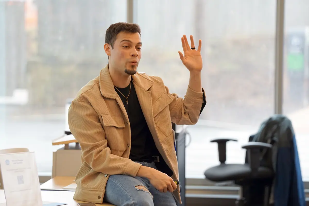 A young man in a tan jacket raises his left hand while sitting on the edge of a desk and speaking in a classroom.
