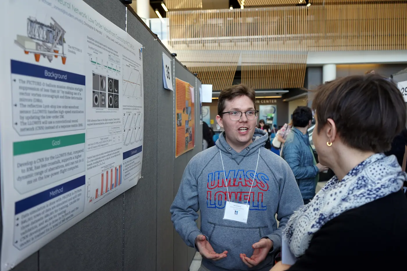 A young man in glasses and a hoodie talks about his research poster with a woman in the lobby of a building.