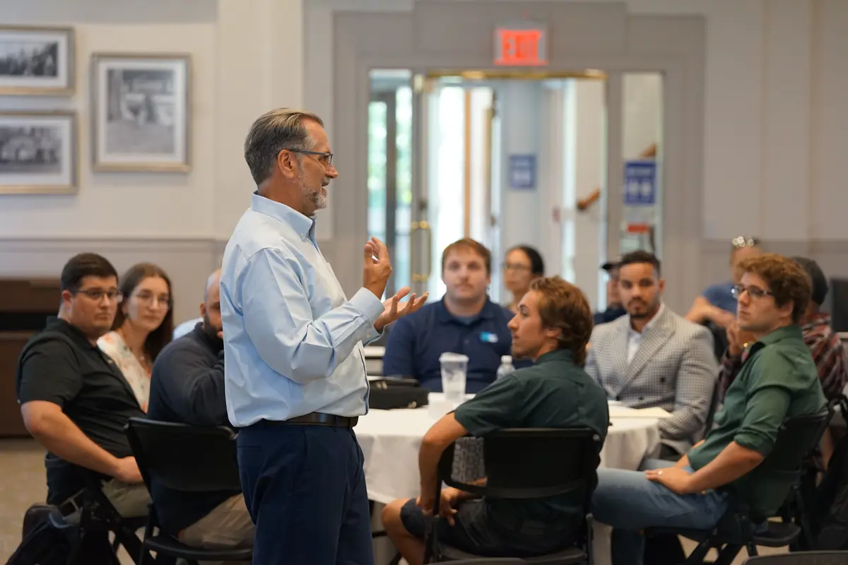 A man in a dress shirt and glasses speaks to a group of people sitting at a round table in a room.