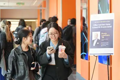 A man in a dark sweater gestures with his hands while standing in the middle of a classroom where students are seated around him.