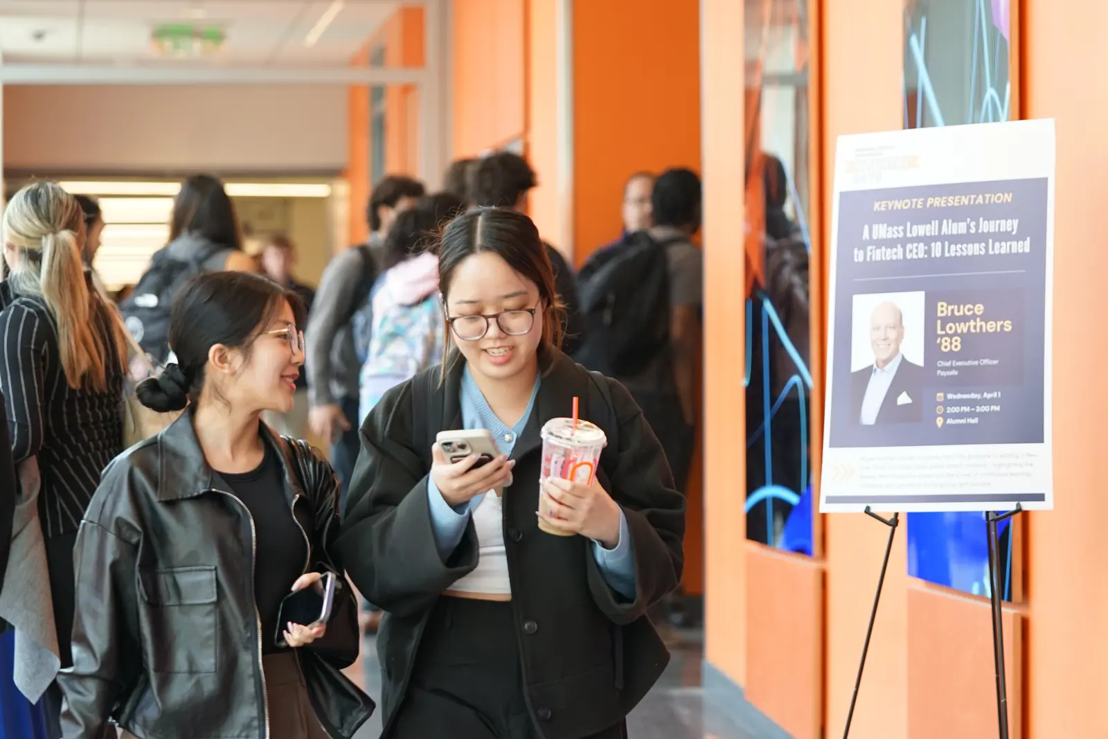 Two college students walk down a hallway while chatting. One is looking at her phone while holding a drink. 
