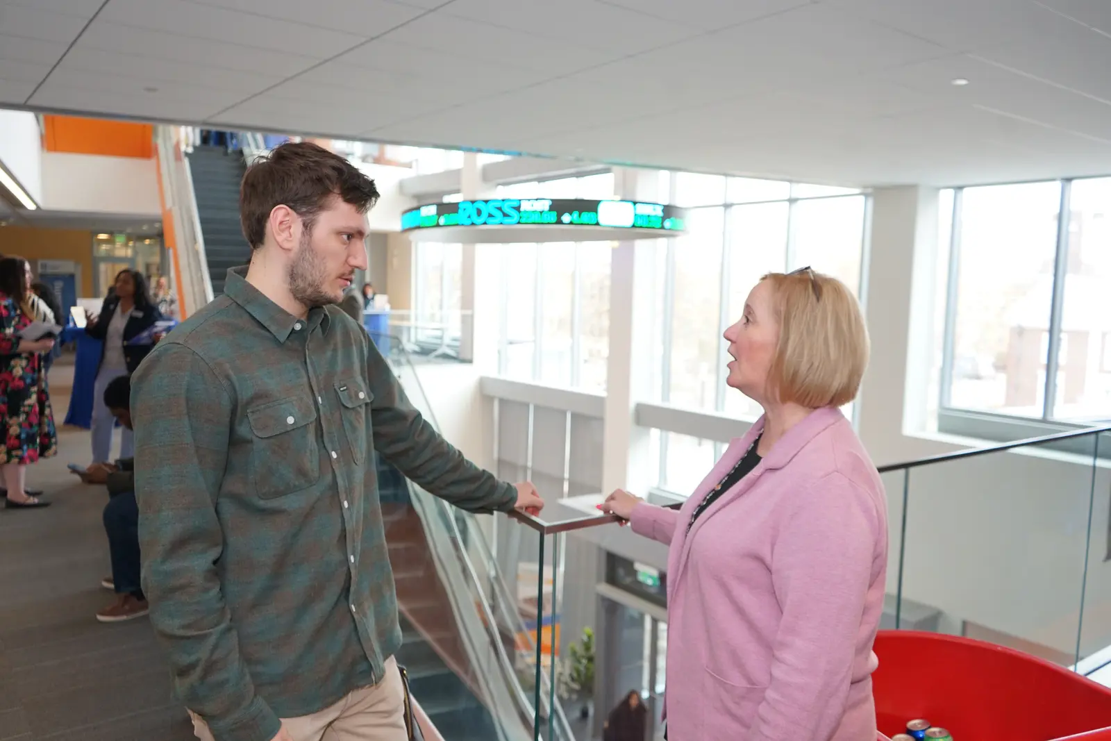 A young man in a green shirt talks to a woman wearing pink while standing along the glass railing inside a building. 