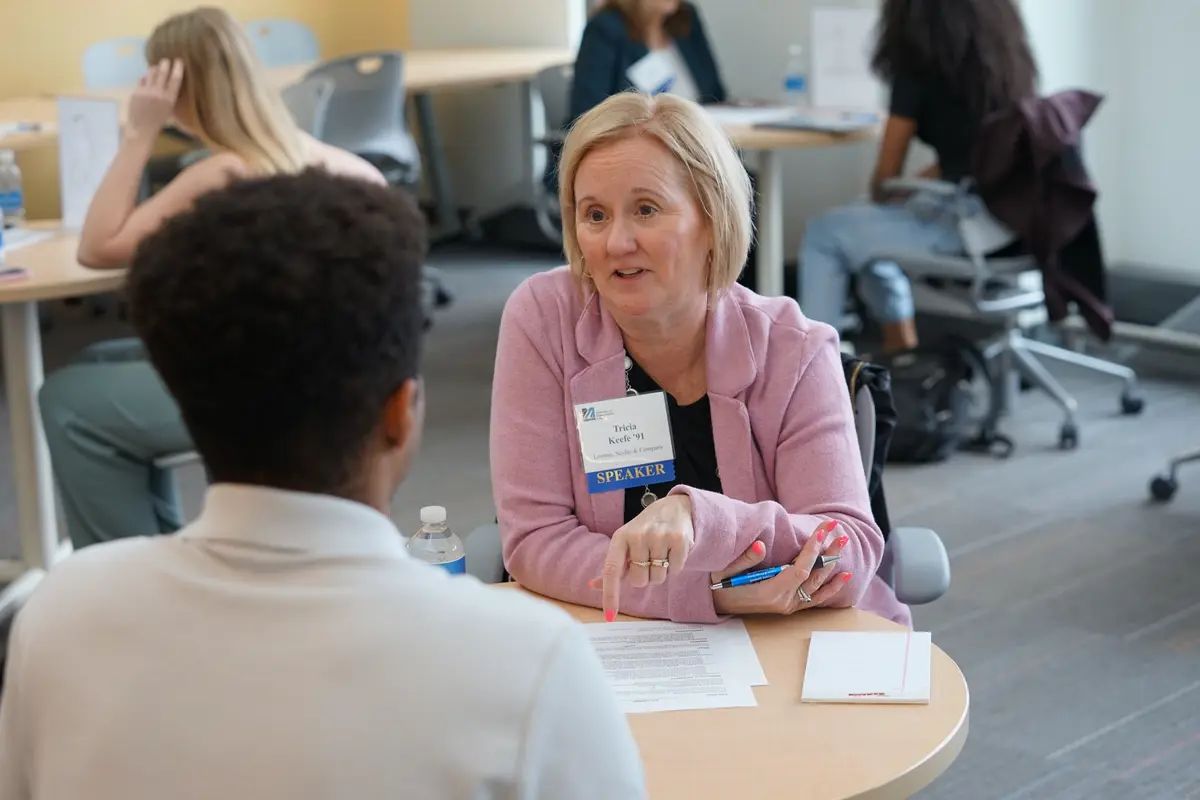 A woman in a pink blazer talks to a college student while seated at a table with papers on it.