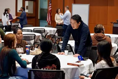 A man in a dark hoodie points to a dry erase board while talking to students.