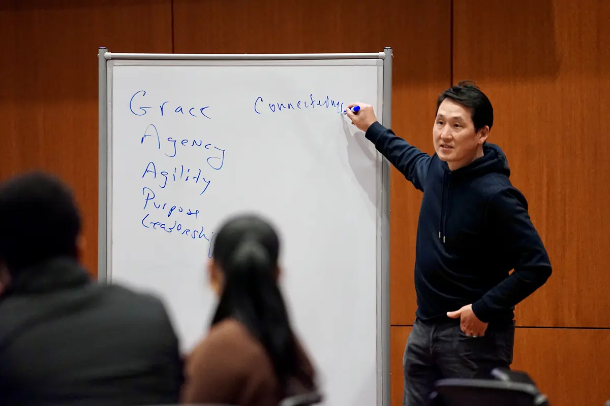 A man in a dark hoodie points to a dry erase board while talking to students.