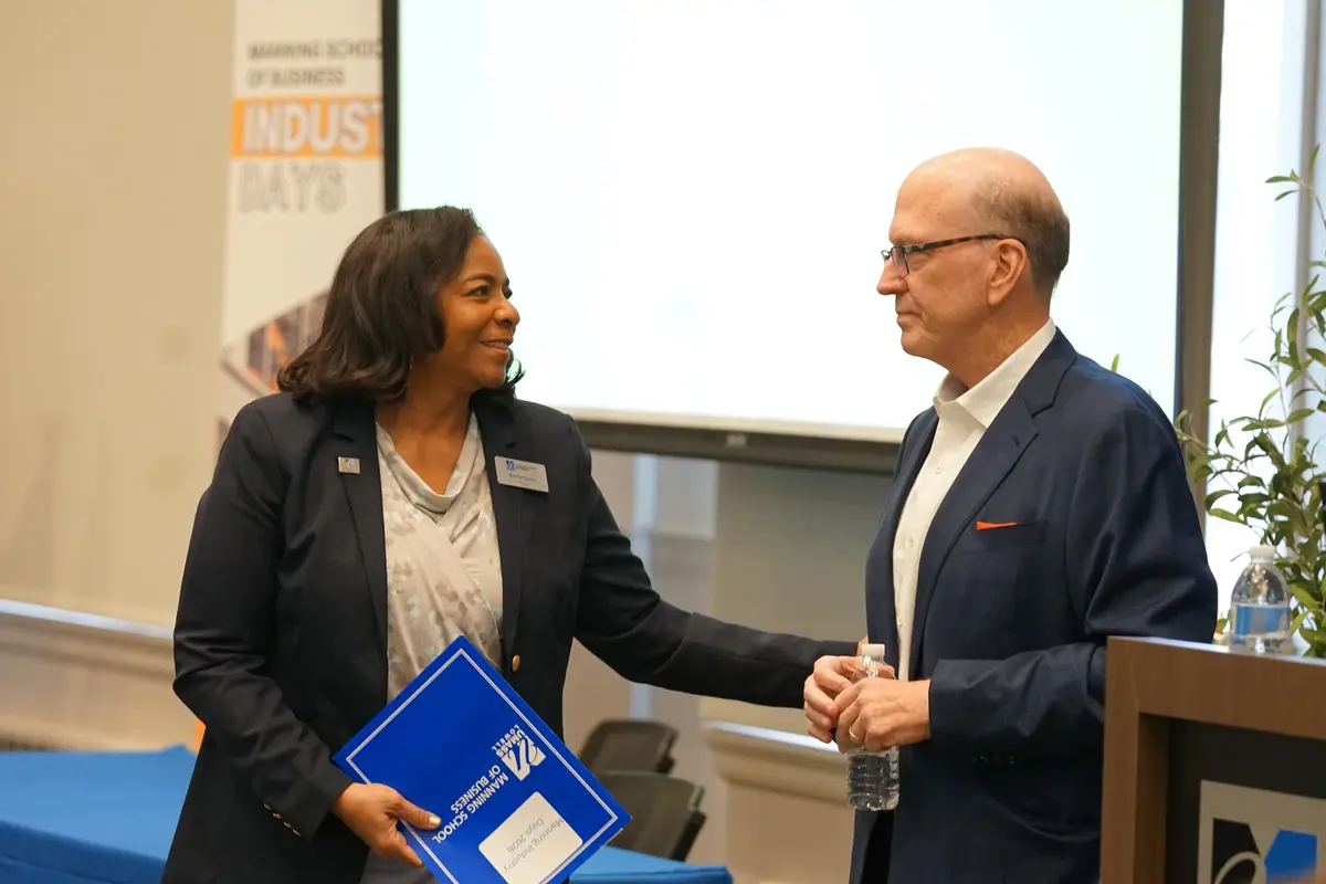 A woman in a blazer smiles while extending her arm toward a man in a suit that is standing next to a lecturn.
