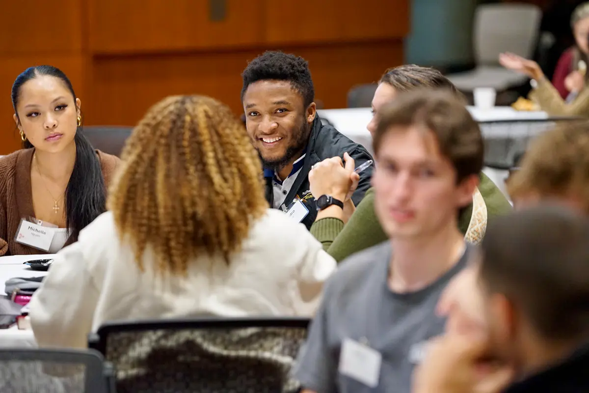 A young man smiles while listening to a person talk seated at a table.