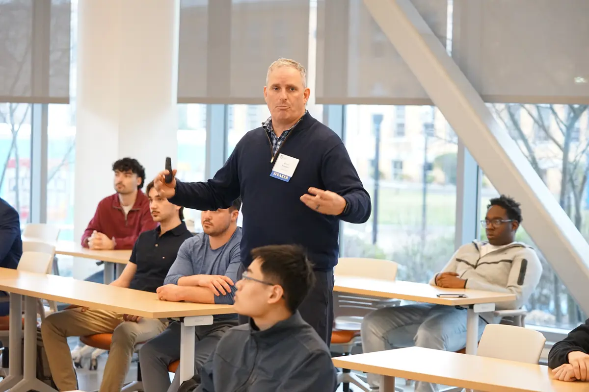 A man in a dark sweater gestures with his hands while standing in the middle of a classroom where students are seated around him.