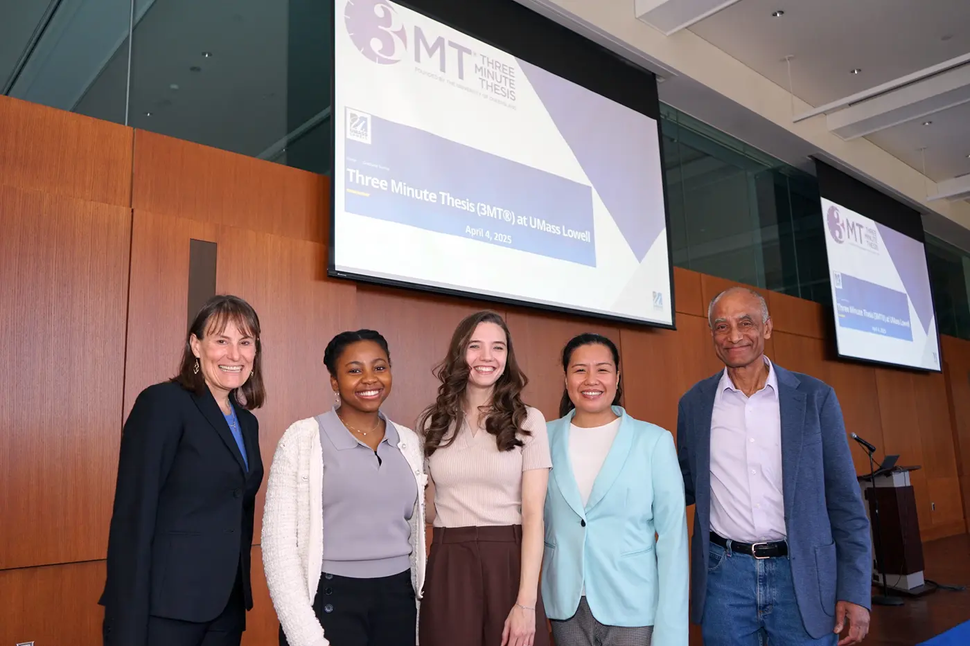 Five people stand in front of a projection screen in a large room and pose for a photo.