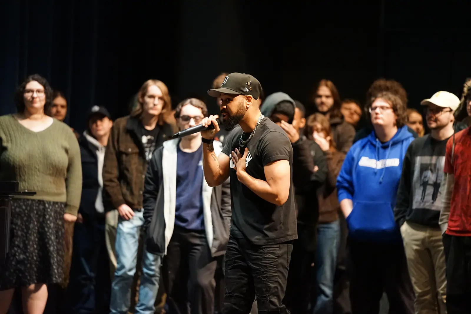 A man in a black hat holds a microphone while talking to a group of college students on a stage.