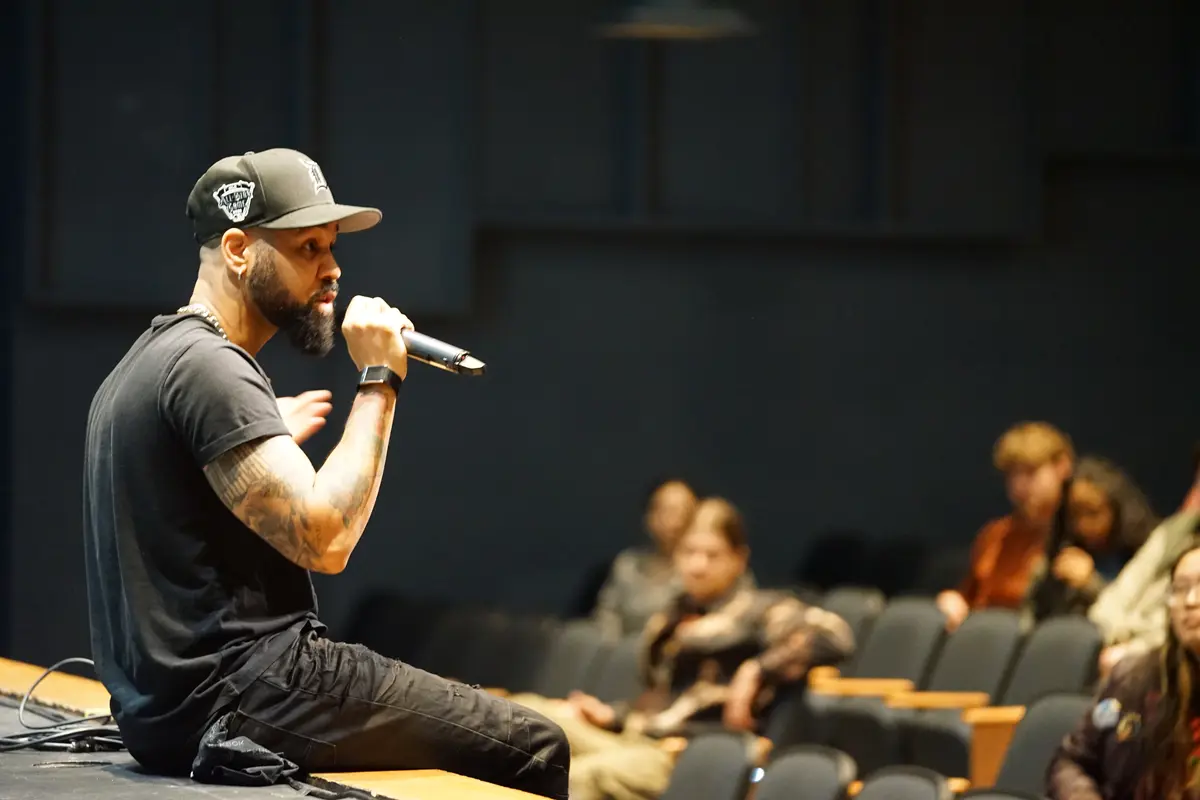 A man in a black T shirt and black baseball cap sits on a stage and talks to students with a microphone.