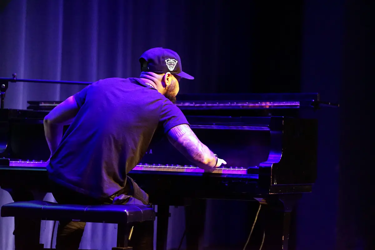 A man in a black T shirt and cap plays a grand piano on a stage.
