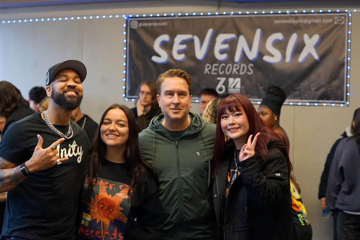 Two men and two women pose for a photo in a room with a poster on the wall behind them that reads SEVENSIX.