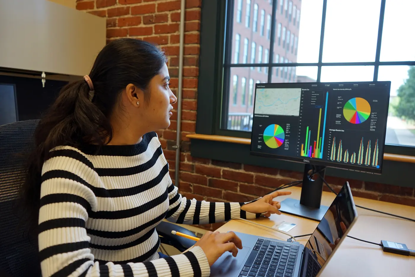 A person looks at a monitor while working on a laptop at a desk near a window.