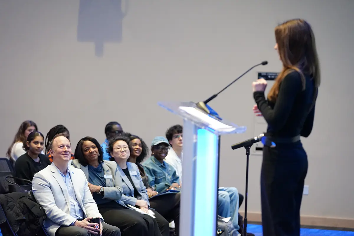 A young woman dressed in black makes a speech at a lecturn while people watch from the audience.