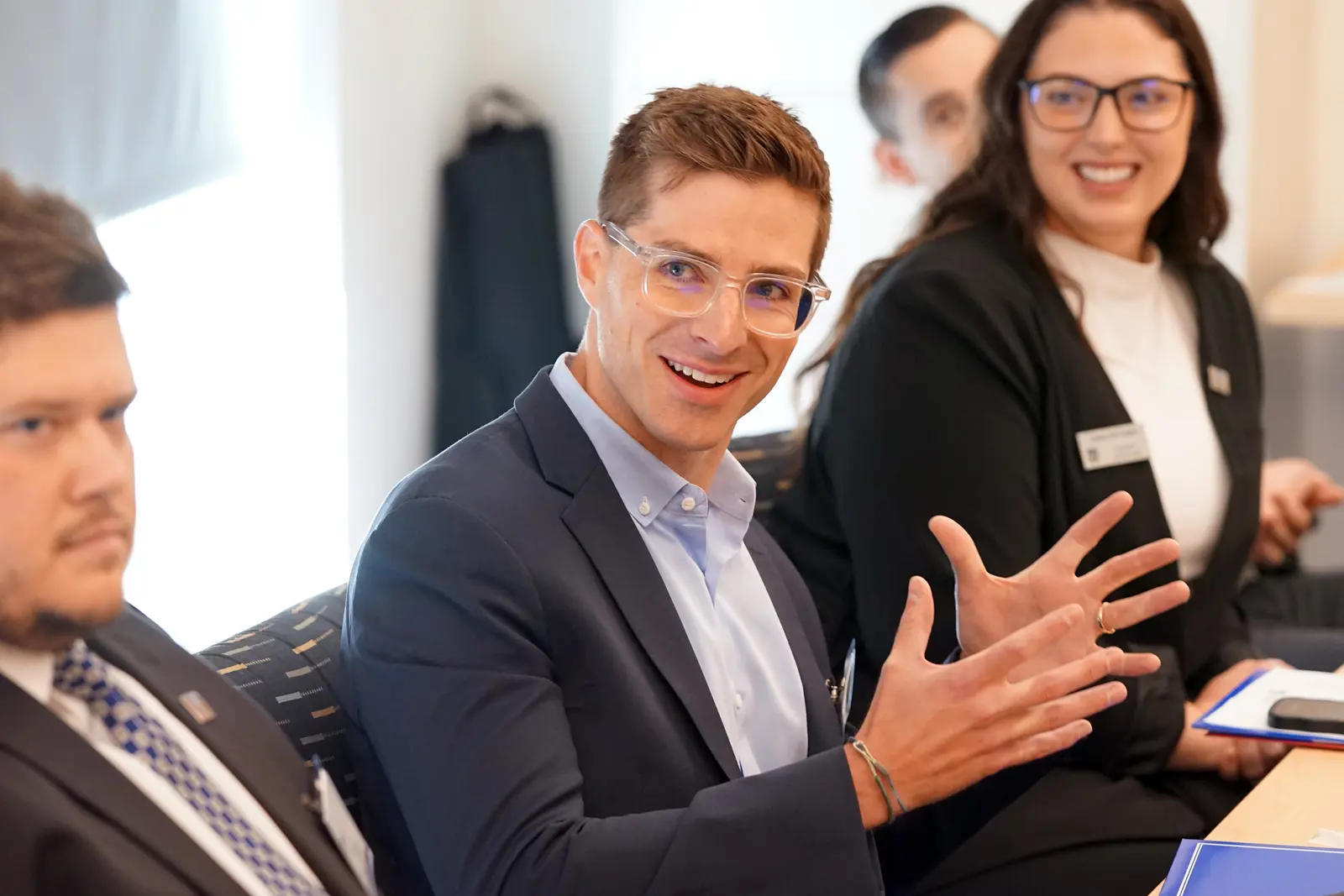 A man in a suit and glasses gestures with his hands while sitting at a table with other people.