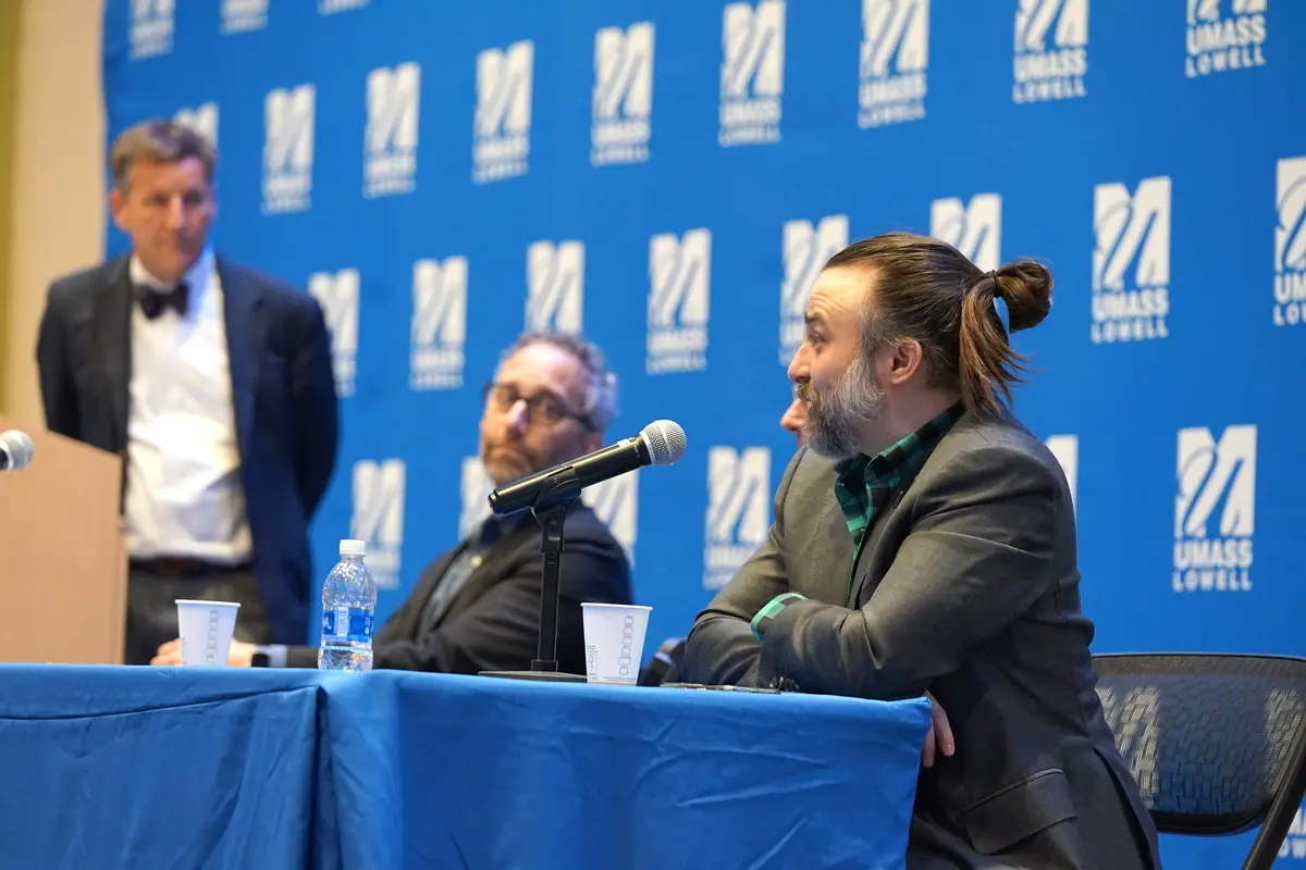 A man in a blazer leans on a table and speaks into a microphone while two other men look on.