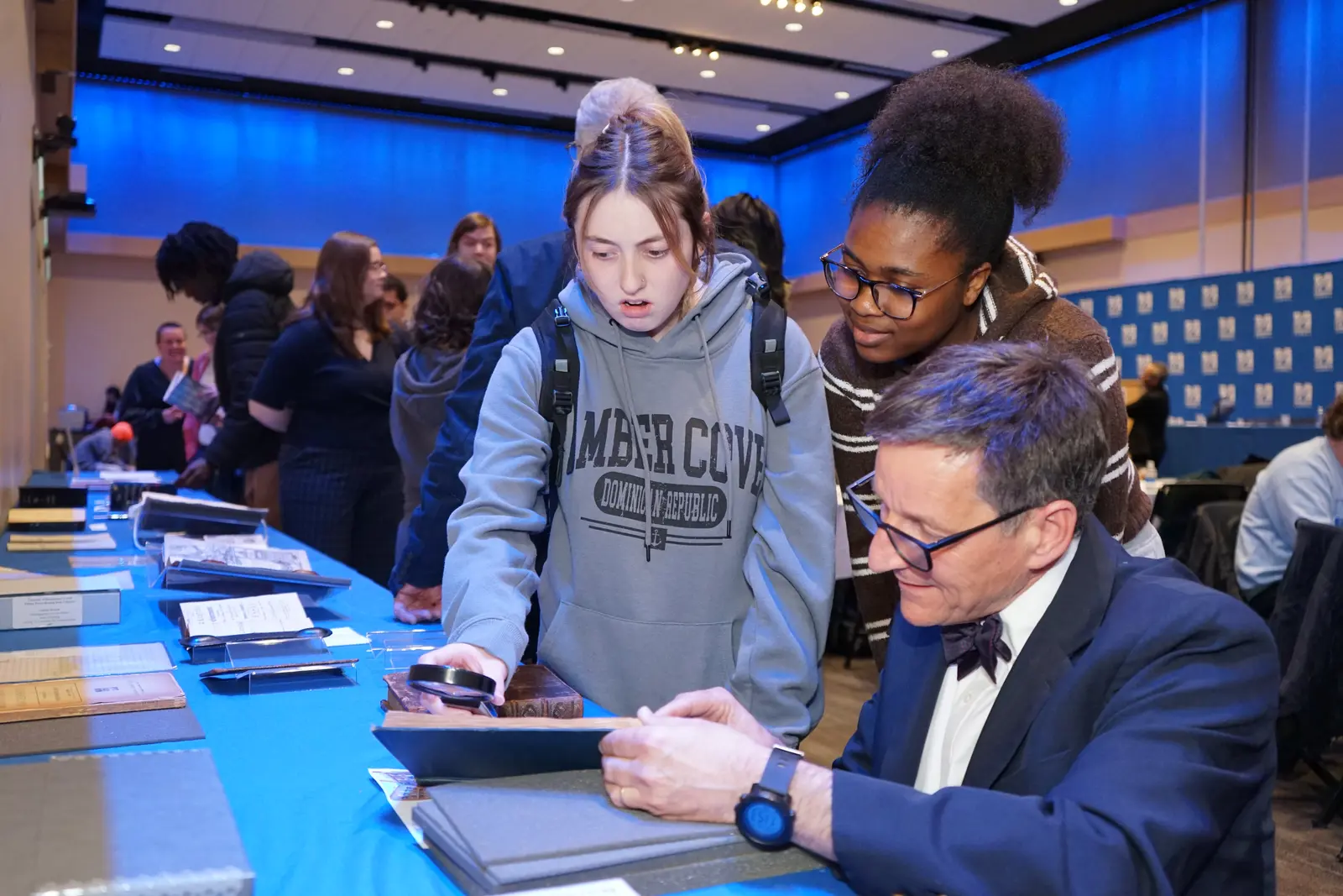 Two college students and a professor look at a book that's on a table in a ballroom.