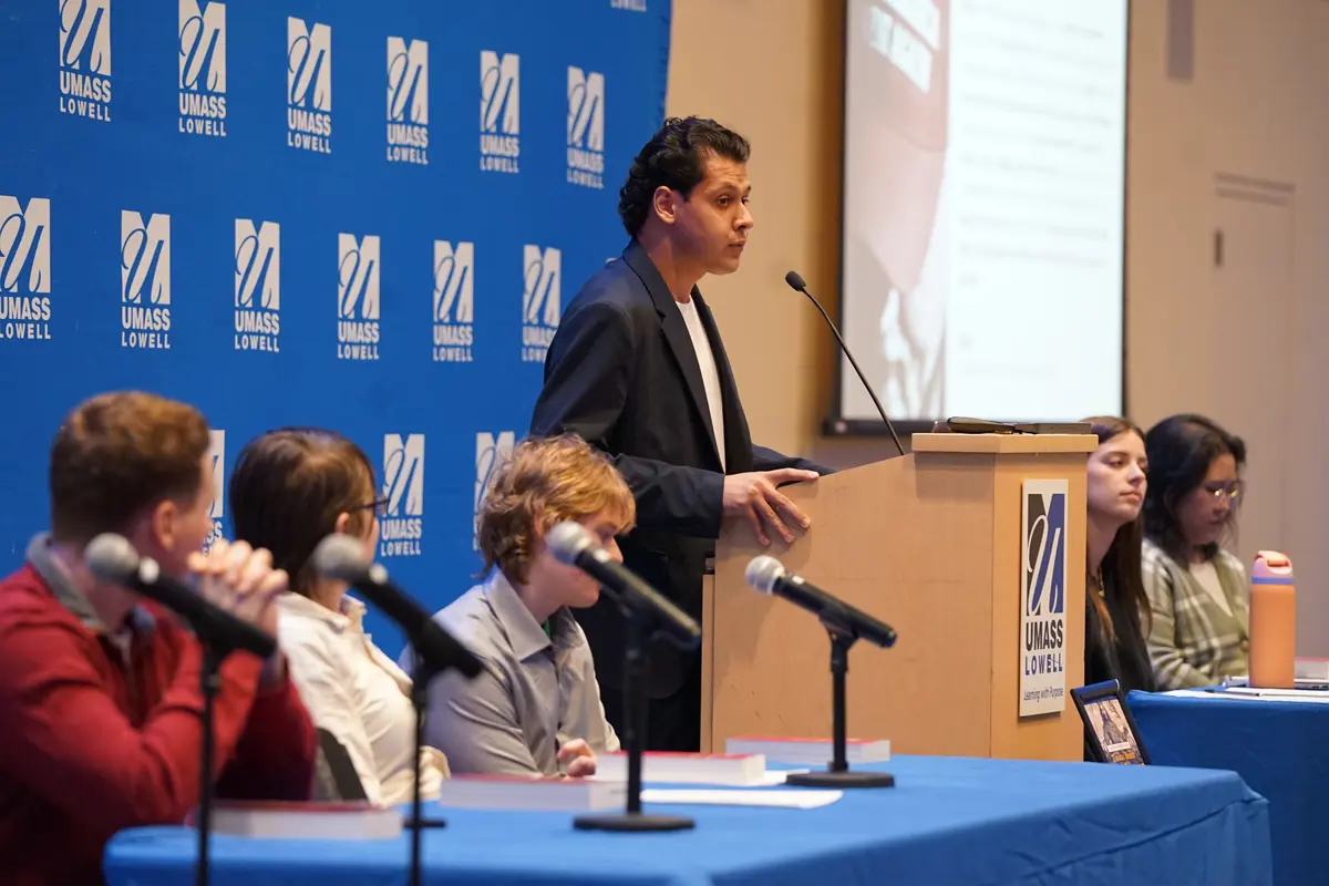 A young man in a blazer speaks at a lecturn while five other college students look on from the dais.
