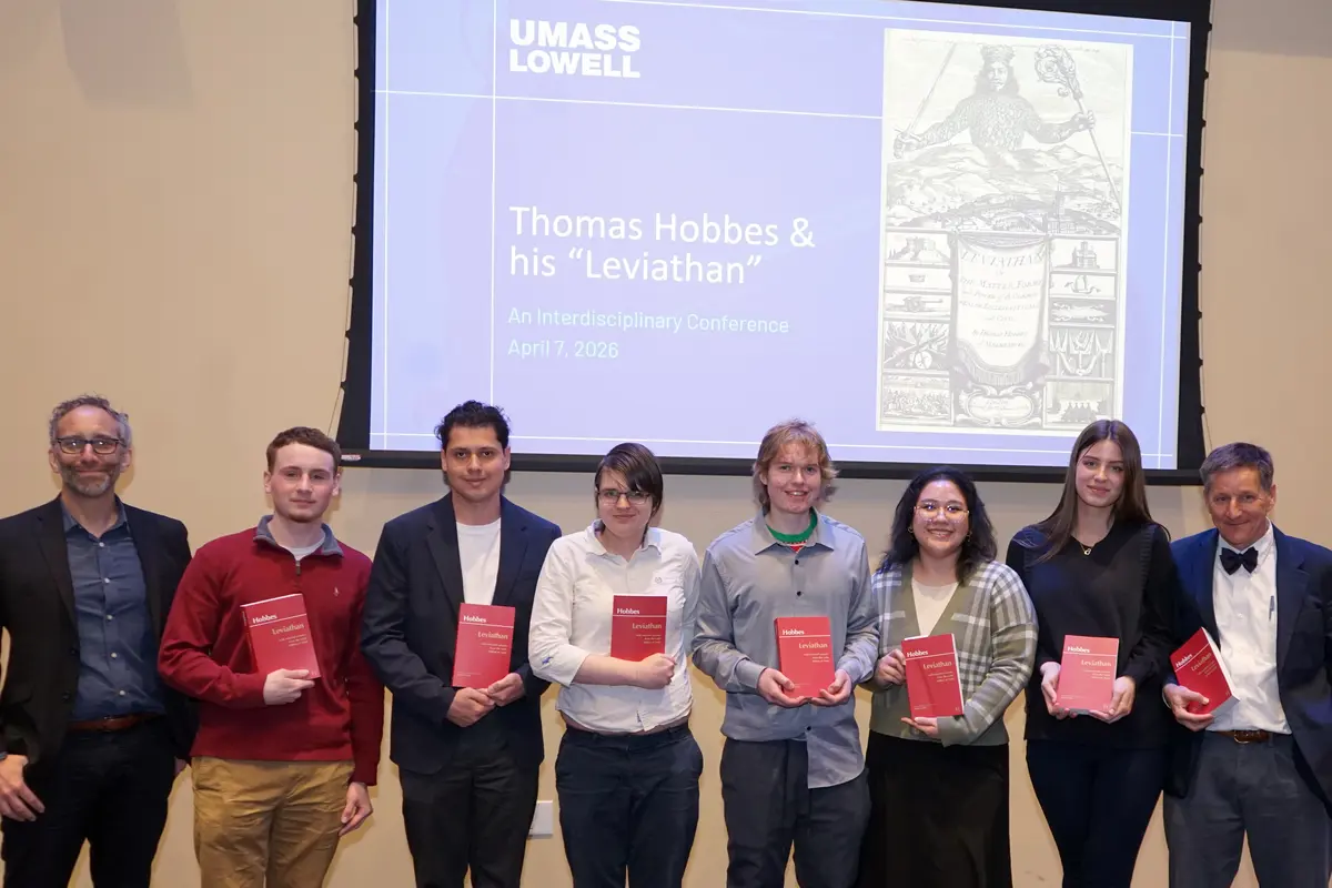 Two men and six college students pose for a photo in front of a projection screen. Seven of them are holding red books.