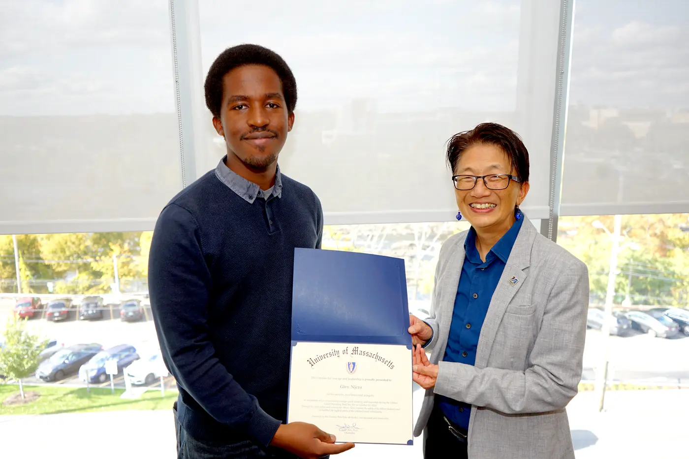 A young man and a woman wearing glasses pose for a photo in an office while holding a certificate.