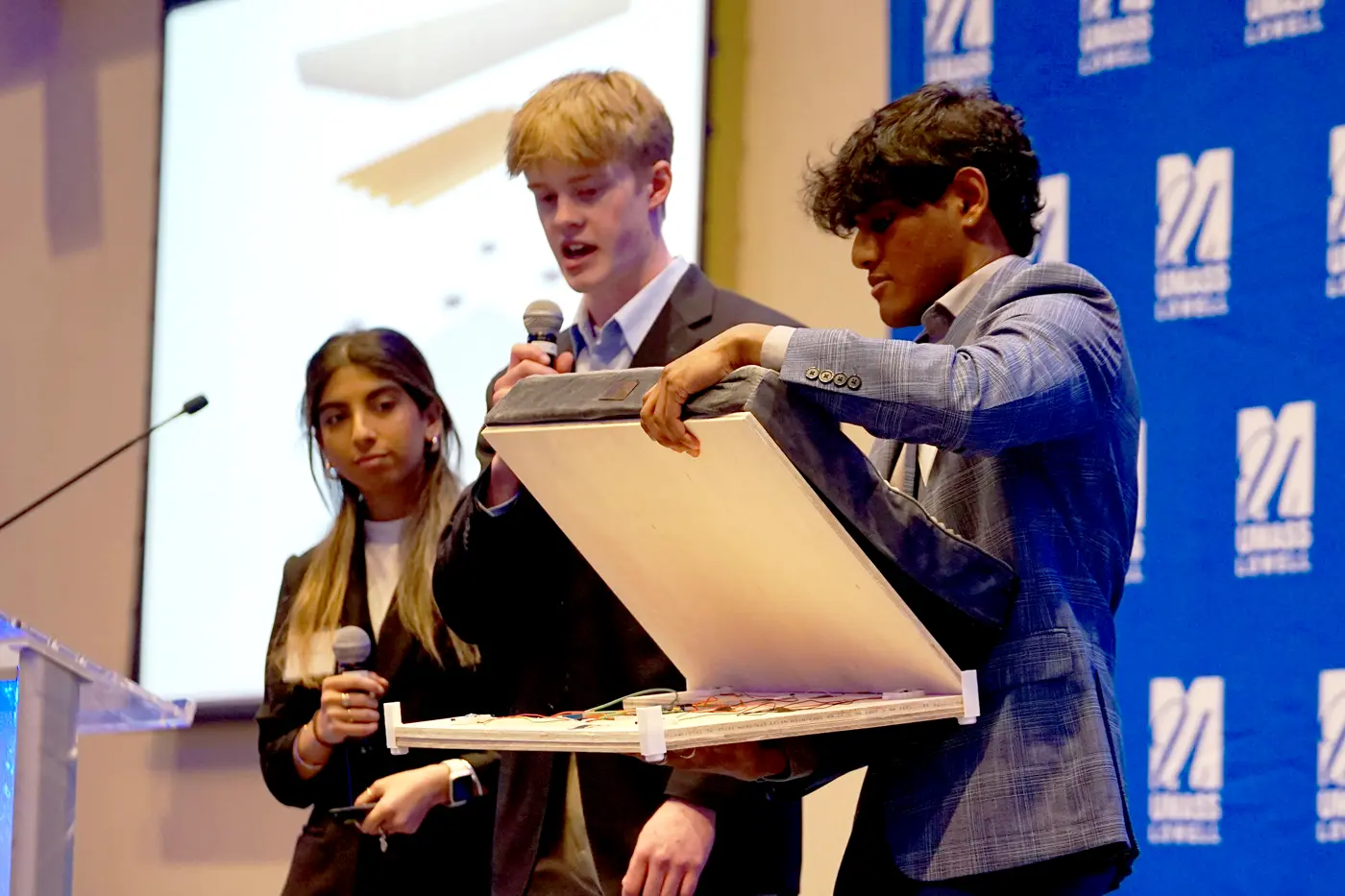 A student opens the top of a smart pet bed while another student speaks into a microphone and another looks on while standing on a stage.