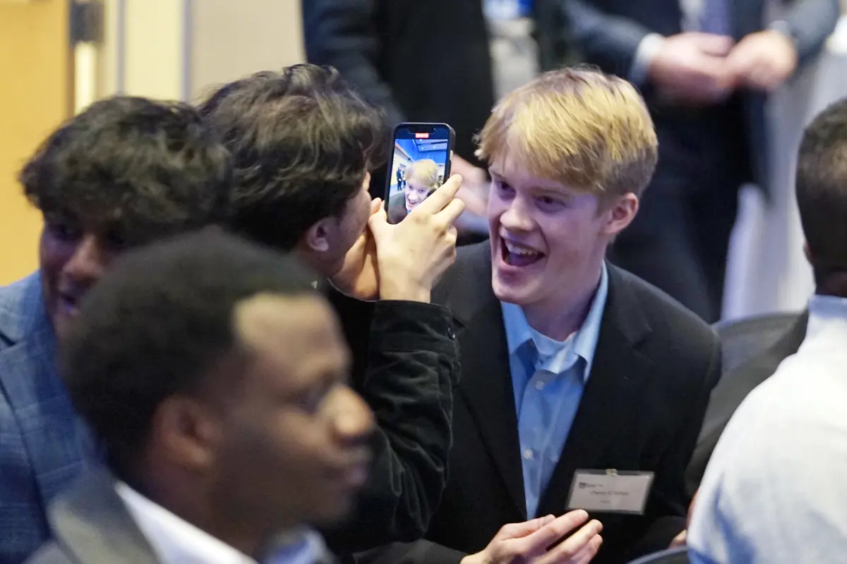 A young man in a blazer smiles while being recorded on a phone by another person while seated.