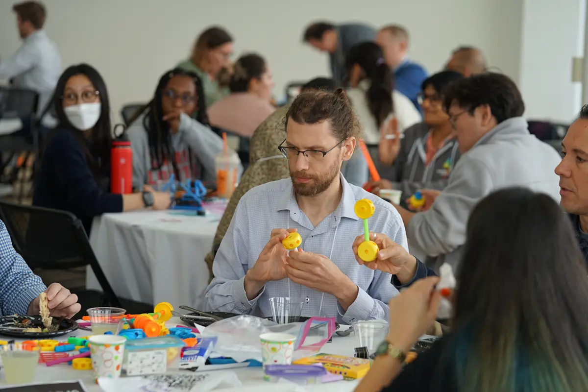 A man holds a tinker toy while seated at a table with other people.