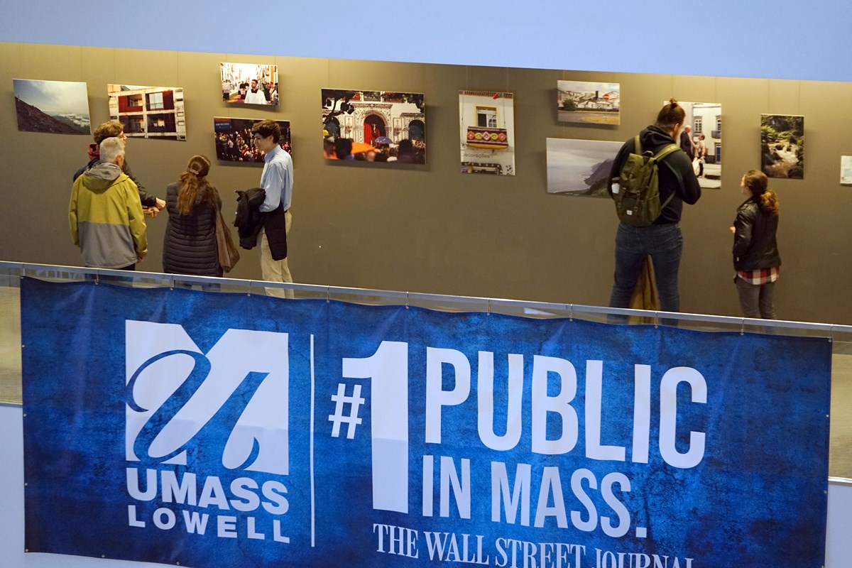 People look at a photo exhibit in a hallway. There is a blue banner hanging on the railing.