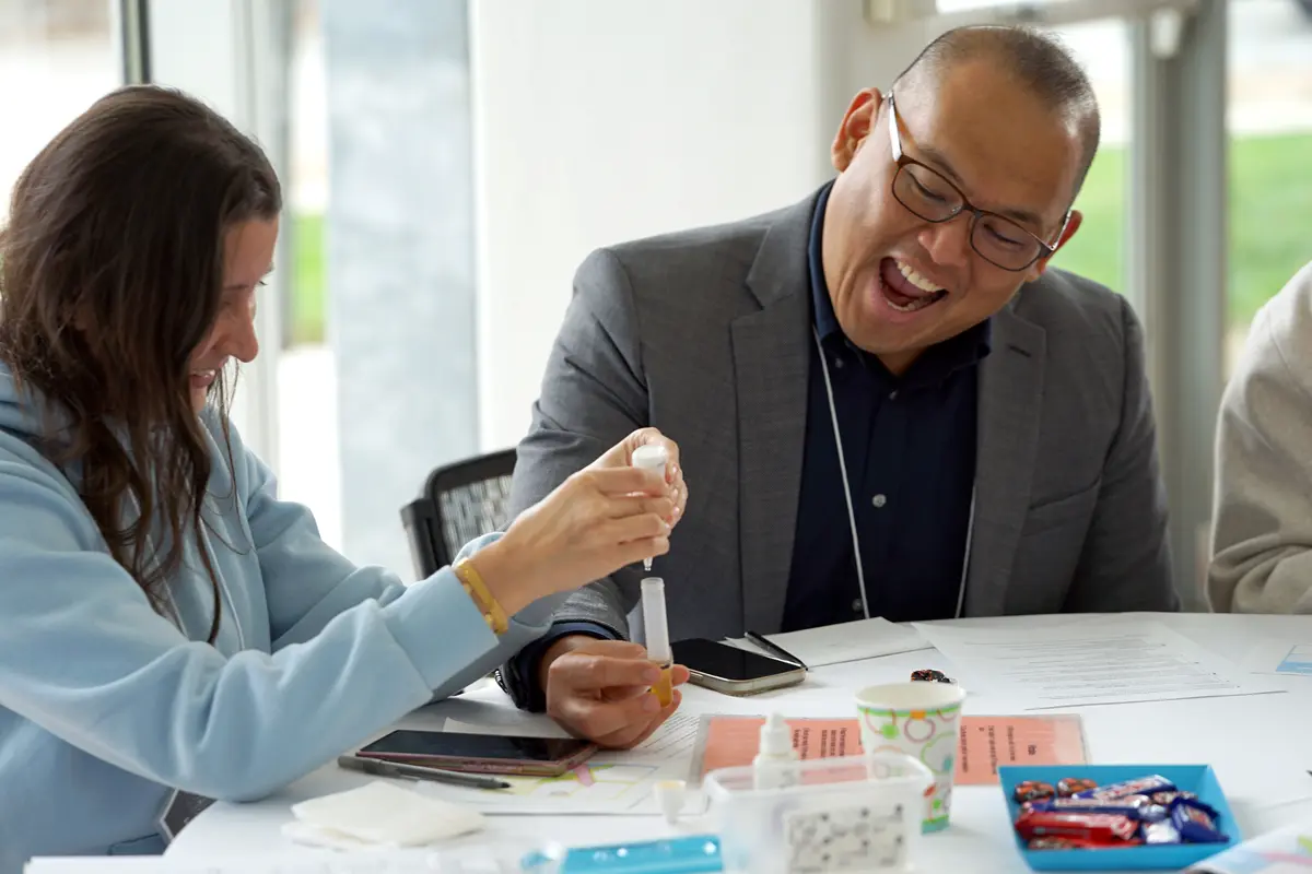 A man in glasses smiles while a woman drops water into a vial while seated at a table.