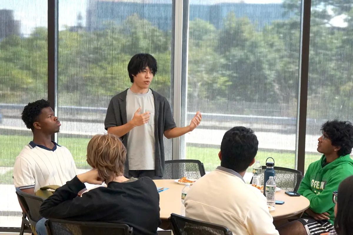A young man with dark hair gestures with his hands while standing at a table of students and speaking.