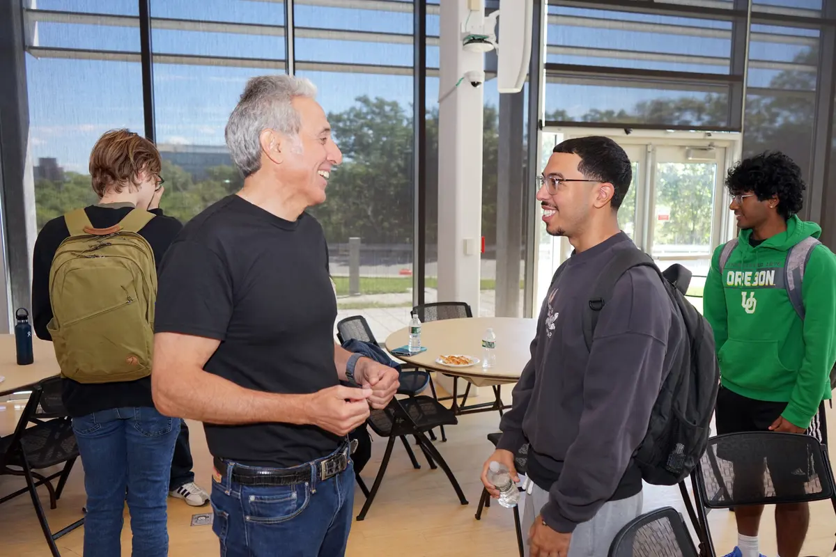 A man in a black T shirt smiles while taking to a college student wearing glasses and a backpack.