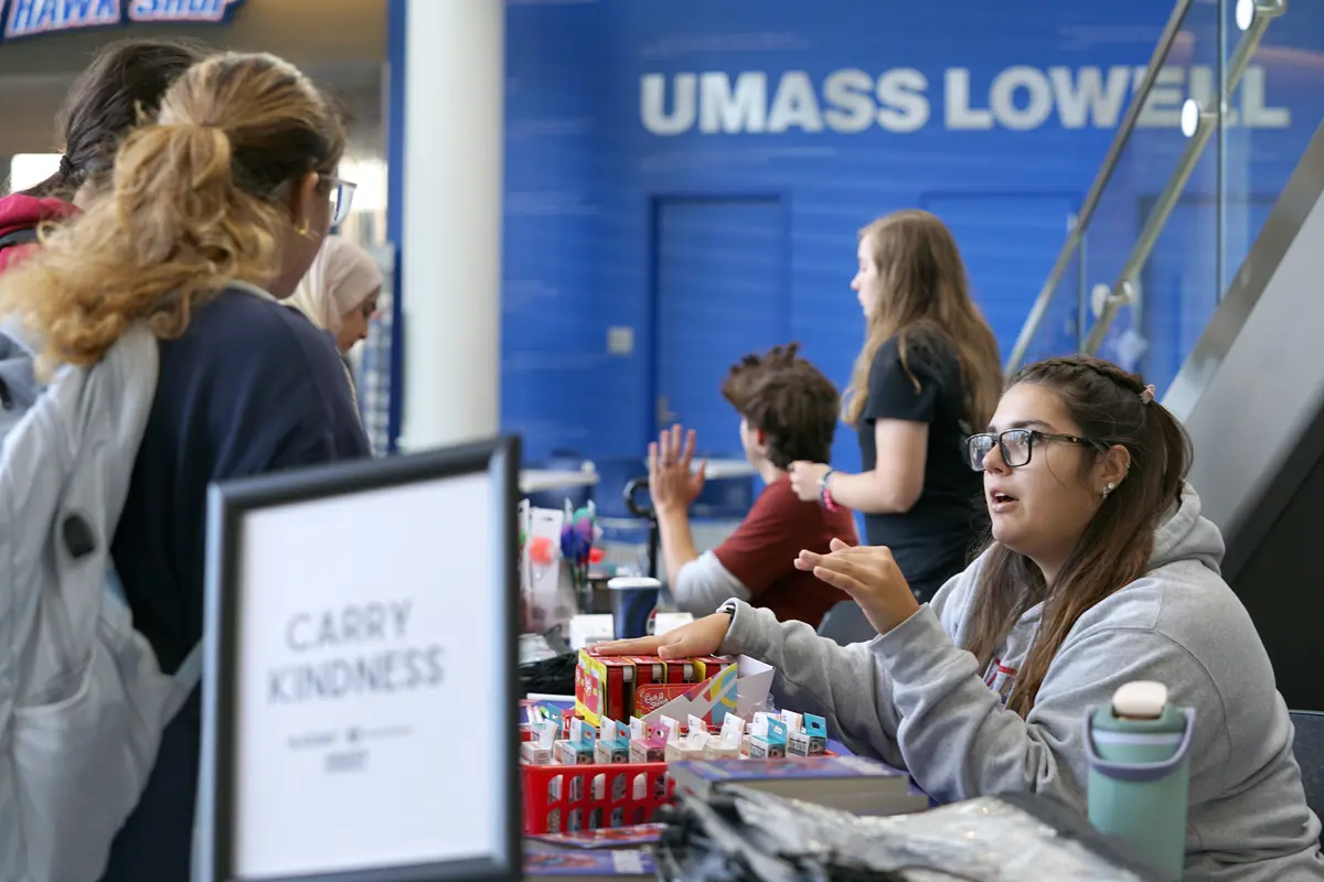 A student in a gray hoodie and glasses sitting a table talks to people in a building lobby.