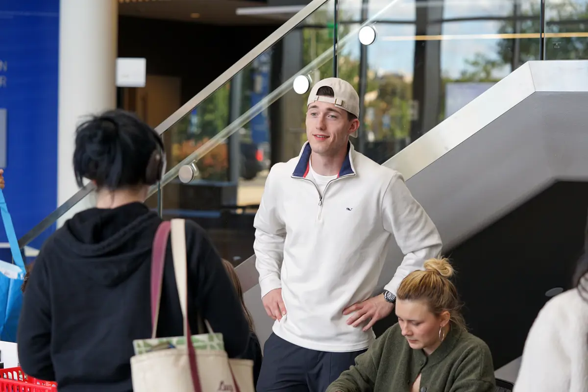 A smiling student stands with his hands on his hips while talking to a person in front of a staircase.