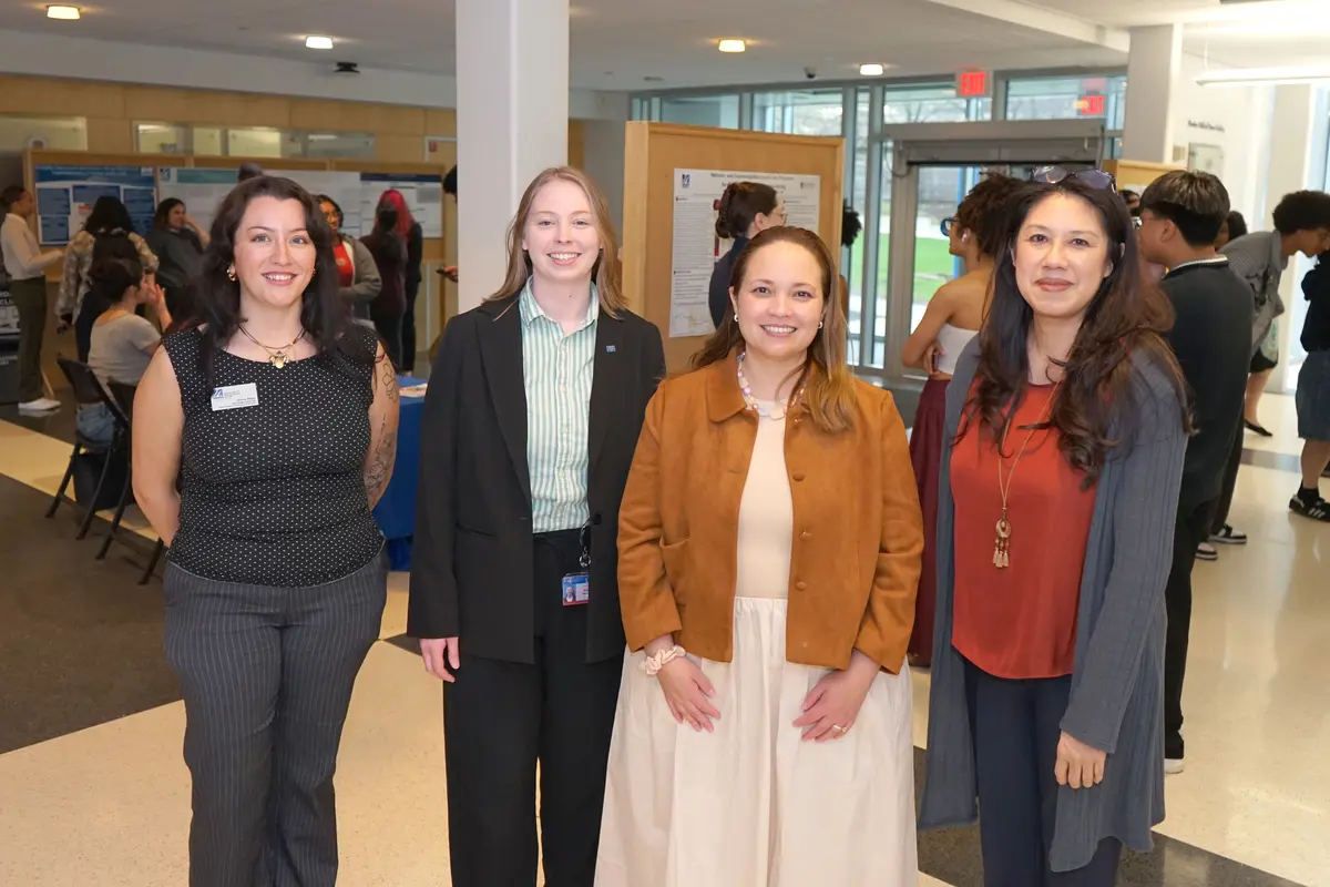 Four women pose for a photo while standing in the lobby of a building where posters are being presented behind them.