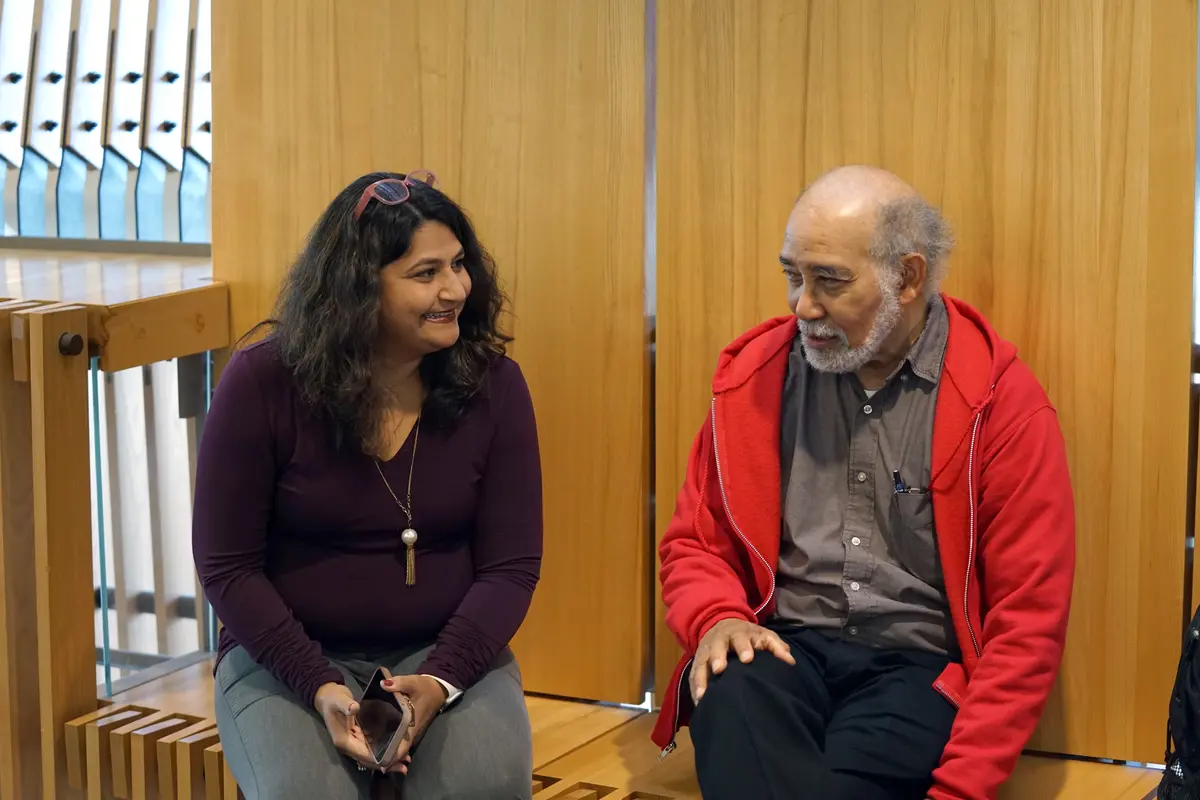 A woman with dark hair smiles while talking to man in a red hoodie while they are sitting on a wood bench indoors.