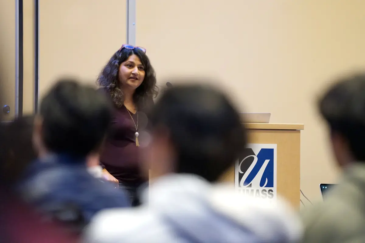 A woman with dark hair stands next to a podium and speaks to people seated in a room.