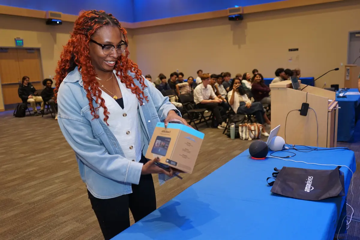 A young woman in glasses holds an Amazon device in a box in front of a table with a blue tablecloth.