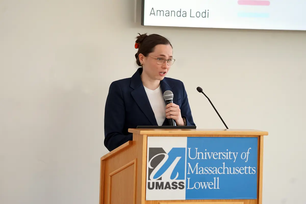 A woman in glasses holds a microphone while speaking at a podium.