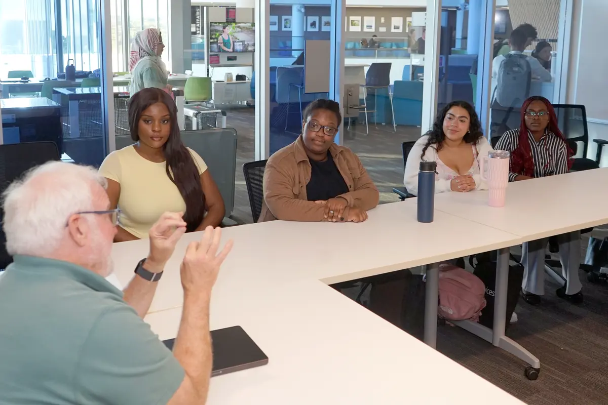 A man gestures with his hands while talking to four young women while sitting around a table in a conference room.