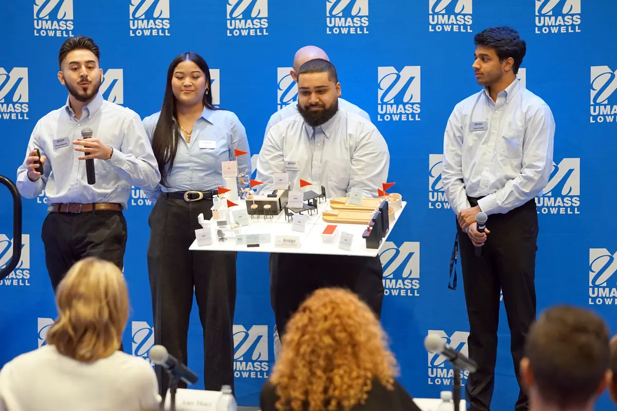 A young man holds a scale model of an entertainment center while standing on a stage in front of other people. 