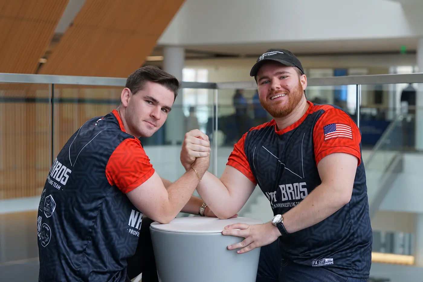 Two young men in matching shirts arm wrestle while posing for a photo in a lobby.