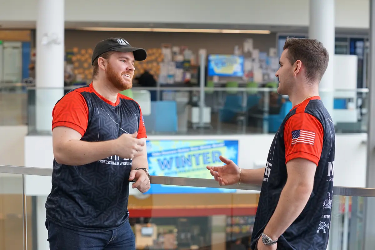 Two young men in matching shirts talk while standing on a balcony in a lobby.