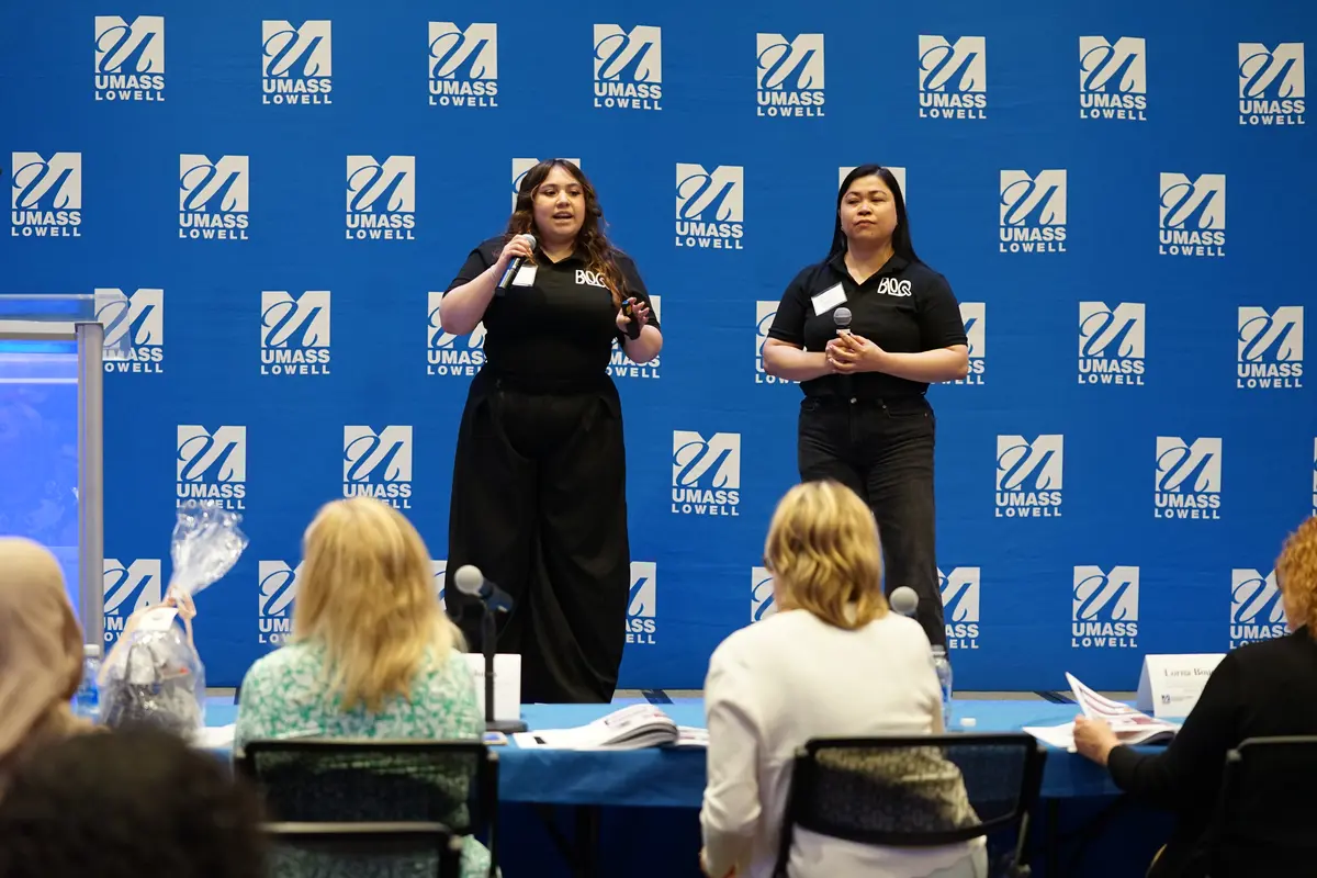 Two young women stand on a stage and speak to a row of people seated at a table.