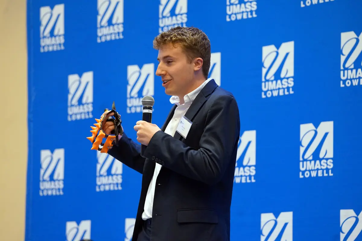 A young man in a blazer holds an orange spiked device while speaking into a microphone.