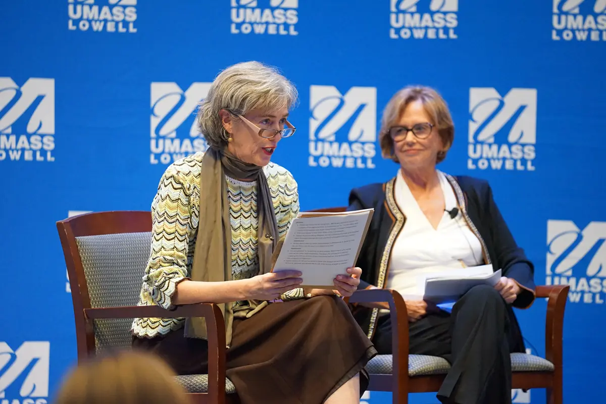 A woman with glasses speaks while sitting next to another woman on a stage.