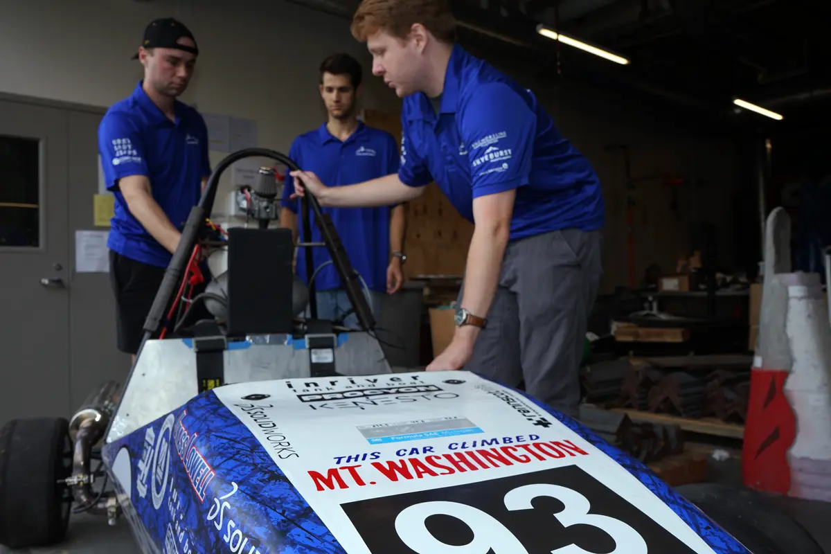 Three young men in matching blue polo shirts stand around a race car in a garage.