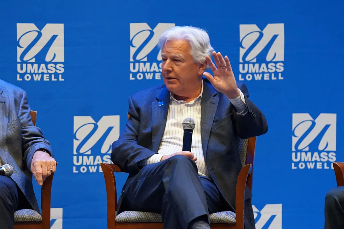 A man in a suit gestures with his hand while holding a microphone and sitting on a stage in front of a blue backdrop.