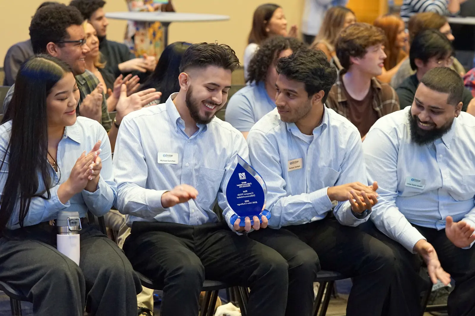Four college students in matching shirts look at a blue glass trophy while sitting in a row in a room.