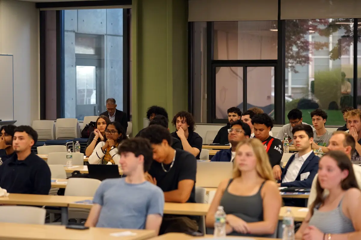 College students listen to a speaker while sitting in rows of desks in a classroom.